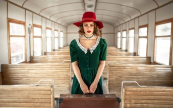 A woman in a vintage-style green dress with lace trim and a red wide-brimmed hat stands in an old-fashioned train carriage. She holds a brown suitcase and looks directly at the camera. The train's wooden benches and large windows create a nostalgic atmosphere.