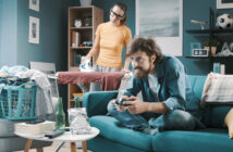 A man with a beard is sitting on a blue couch playing video games with a controller, while a woman is ironing clothes behind him in a living room. The room is cluttered with laundry, a laundry basket, snacks, and drinks, and is decorated with wall art and a bookshelf.
