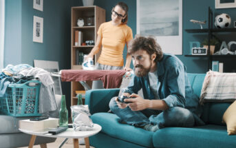 A man with a beard is sitting on a blue couch playing video games with a controller, while a woman is ironing clothes behind him in a living room. The room is cluttered with laundry, a laundry basket, snacks, and drinks, and is decorated with wall art and a bookshelf.