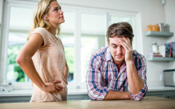 A young woman with blond hair stands in a kitchen with her hands on her hips, looking frustrated. A young man with brown hair wearing a plaid shirt sits at the counter, resting his head on his hand, appearing upset.