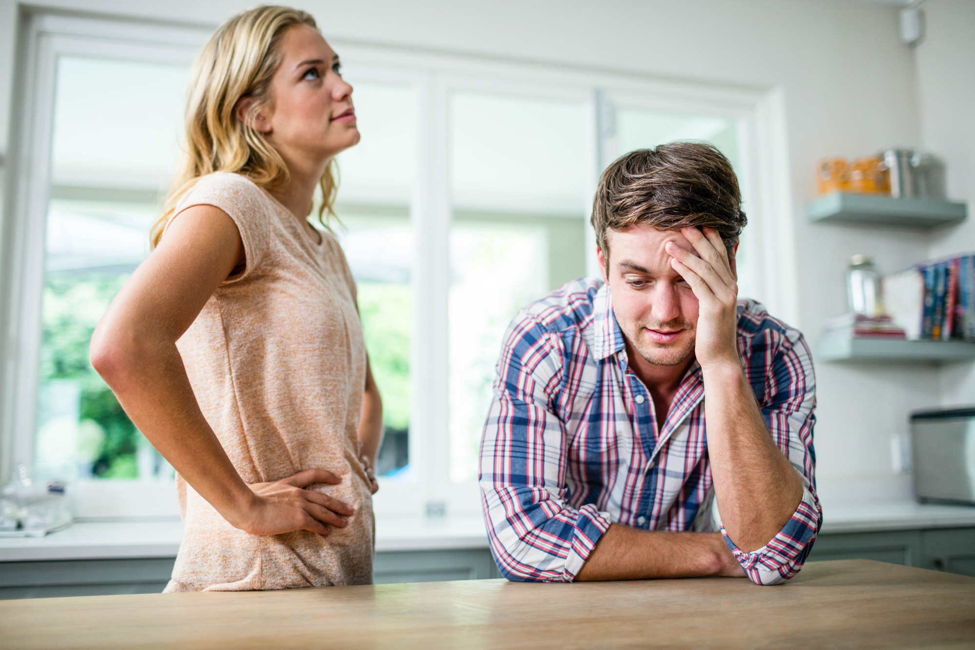 A young woman with blond hair stands in a kitchen with her hands on her hips, looking frustrated. A young man with brown hair wearing a plaid shirt sits at the counter, resting his head on his hand, appearing upset.