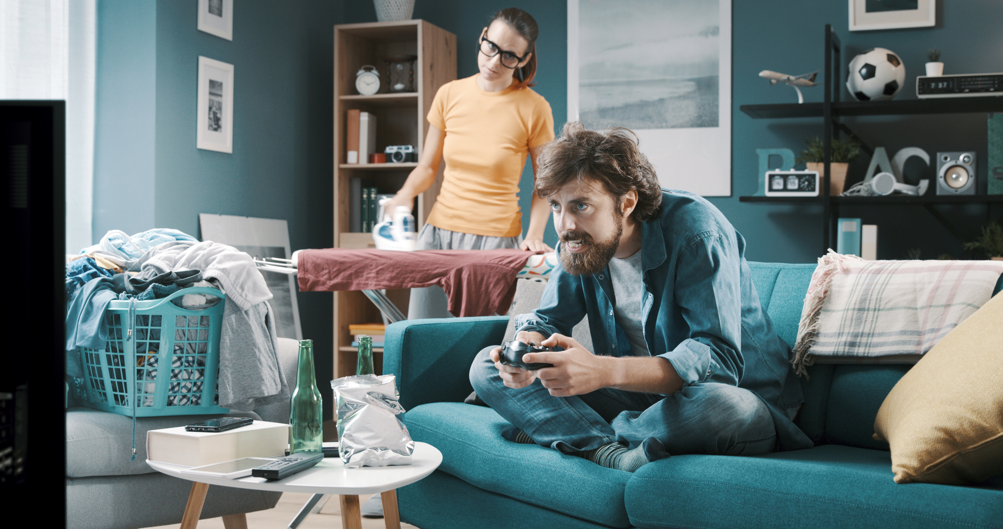 A man with a beard is sitting on a blue couch playing video games with a controller, while a woman is ironing clothes behind him in a living room. The room is cluttered with laundry, a laundry basket, snacks, and drinks, and is decorated with wall art and a bookshelf.