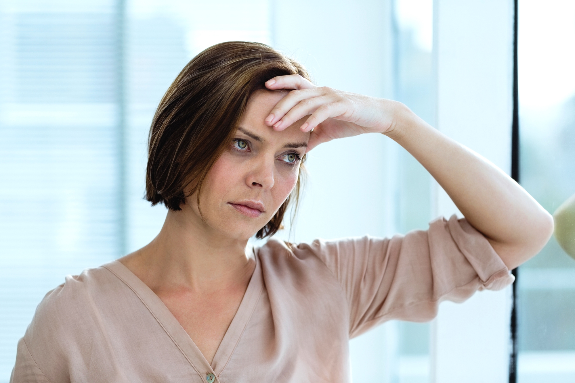 A woman with short brown hair, wearing a light peach blouse, stands by a window. She appears concerned or reflective, resting her forehead on her right hand while leaning slightly to the side. Natural light filters through the window behind her.