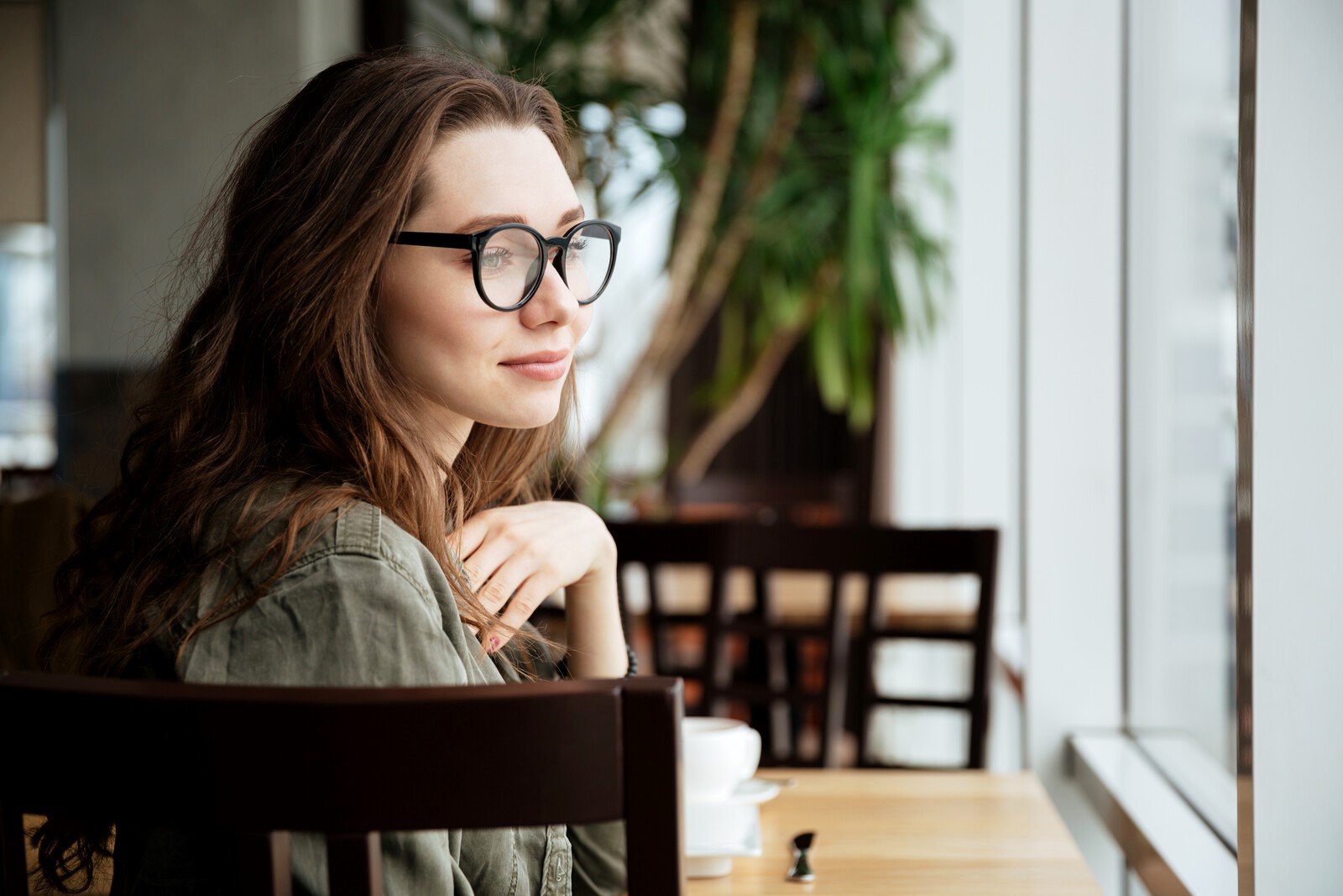 Woman with glasses and long, brown hair, sits at a wooden table in a cafe, looking out of a large window. She wears an olive green jacket and appears to be relaxed and thoughtful. In the background, there are chairs and a tall plant.