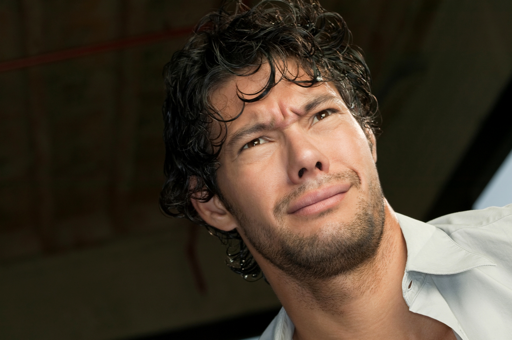 A man with curly dark hair and a light beard is looking off into the distance with a confused or concerned expression. He is wearing a light-colored shirt and is photographed from a low angle. The background is out of focus, suggesting an indoor setting.