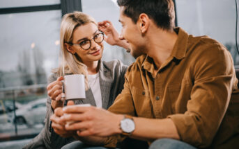 A woman with blonde hair and glasses holds a white mug, adjusting her glasses with her other hand, while looking at a man with dark hair. The man, wearing a brown jacket, holds a cup and smiles at her. They sit on a couch with a large window in the background.