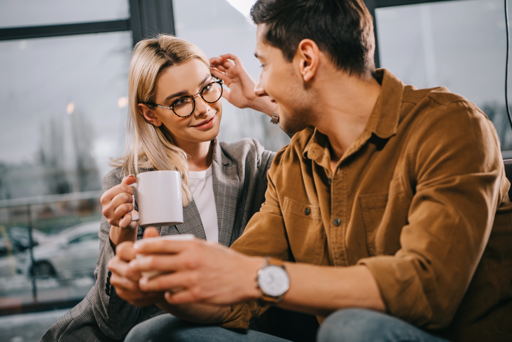 A woman with blonde hair and glasses holds a white mug, adjusting her glasses with her other hand, while looking at a man with dark hair. The man, wearing a brown jacket, holds a cup and smiles at her. They sit on a couch with a large window in the background.