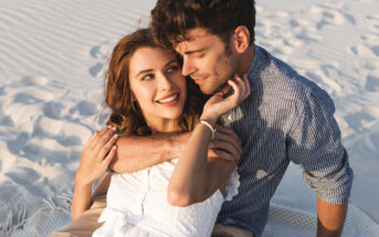 A couple sits closely together on a sandy beach. The woman, wearing a white dress, smiles brightly while looking up at the man, who is in a striped shirt and has his arm around her shoulders. They appear happy and content, surrounded by rippled sand patterns.