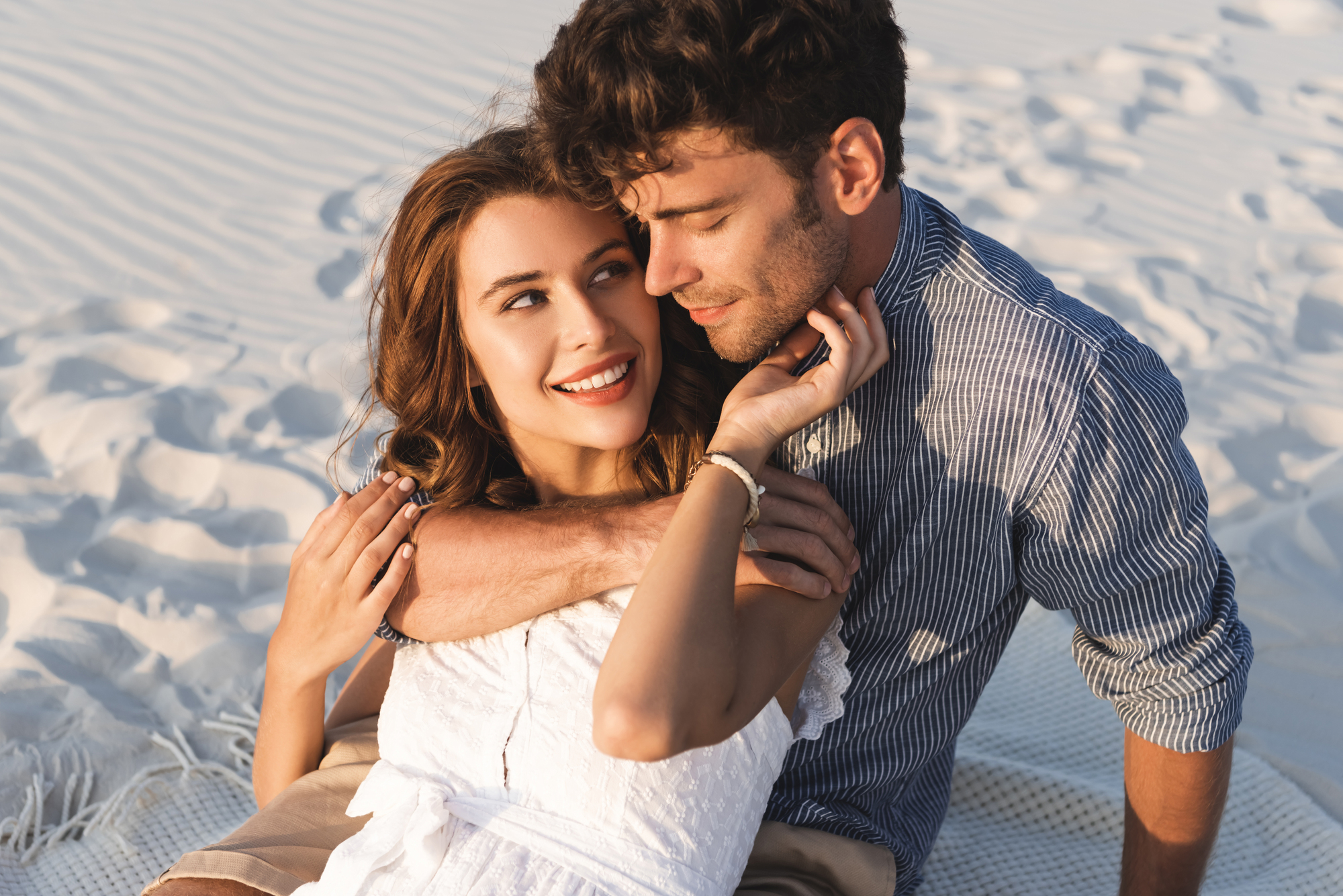 A couple sits closely together on a sandy beach. The woman, wearing a white dress, smiles brightly while looking up at the man, who is in a striped shirt and has his arm around her shoulders. They appear happy and content, surrounded by rippled sand patterns.