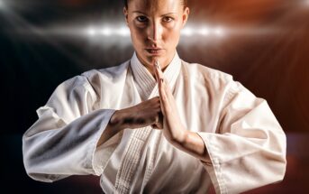 A person dressed in a white martial arts uniform stands with a serious expression, hands held together in a traditional greeting or prayer position. The background is dark with a spotlight illuminating from behind, creating a dramatic effect.