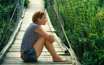 A person with short hair and wearing a striped shirt and denim shorts sits barefoot on a wooden suspension bridge. They are resting their arms on their knees and looking outwards, surrounded by lush green vegetation.