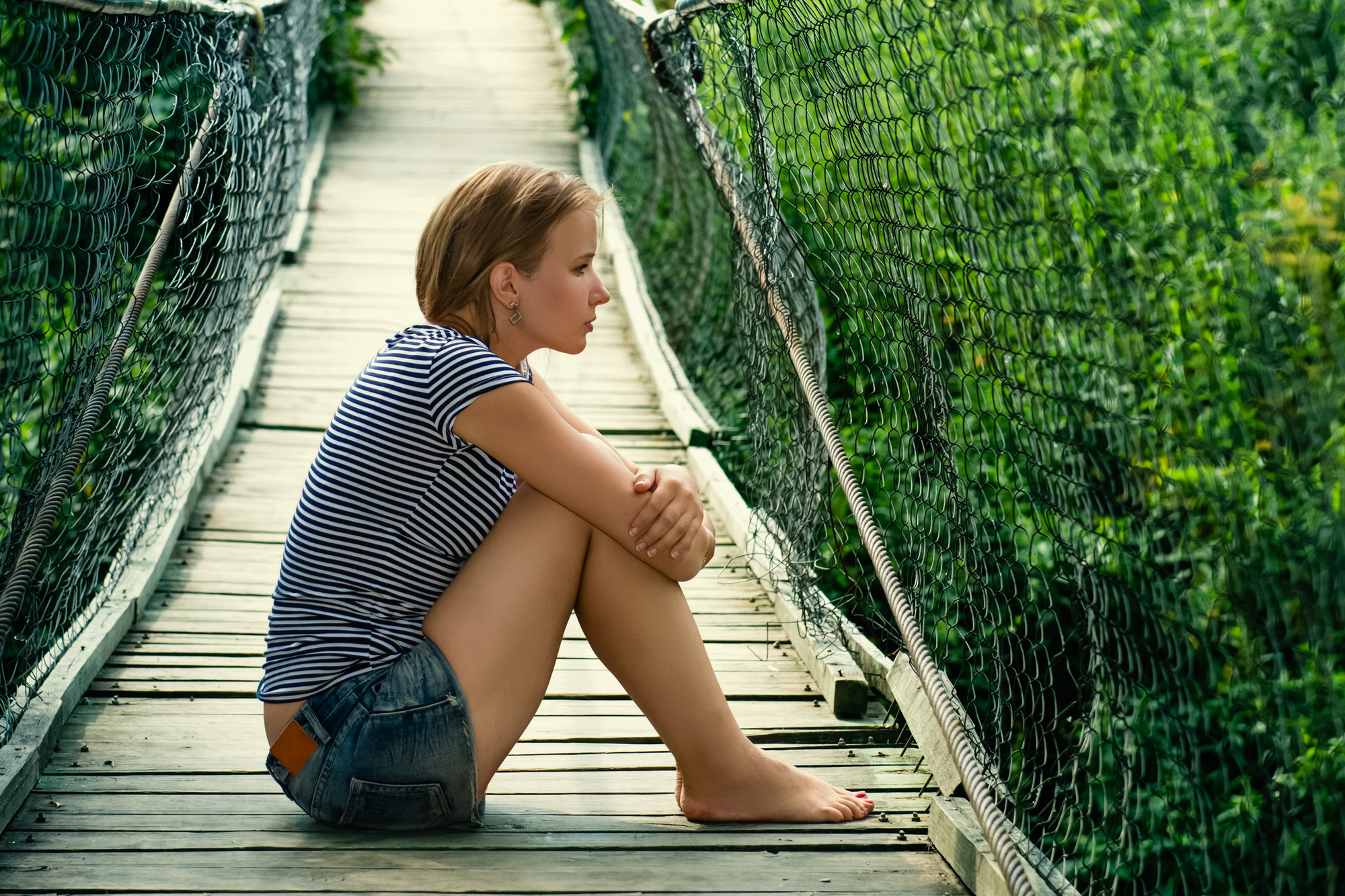 A person with short hair and wearing a striped shirt and denim shorts sits barefoot on a wooden suspension bridge. They are resting their arms on their knees and looking outwards, surrounded by lush green vegetation.