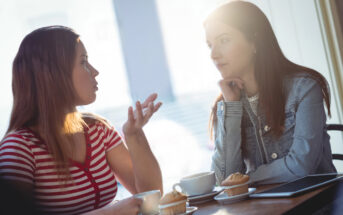 Two women are sitting at a table in a café, engaged in a conversation. One has her hand raised in a gesturing motion, while the other rests her chin on her hand, listening intently. On the table are two coffee cups, saucers, muffins, and a tablet.