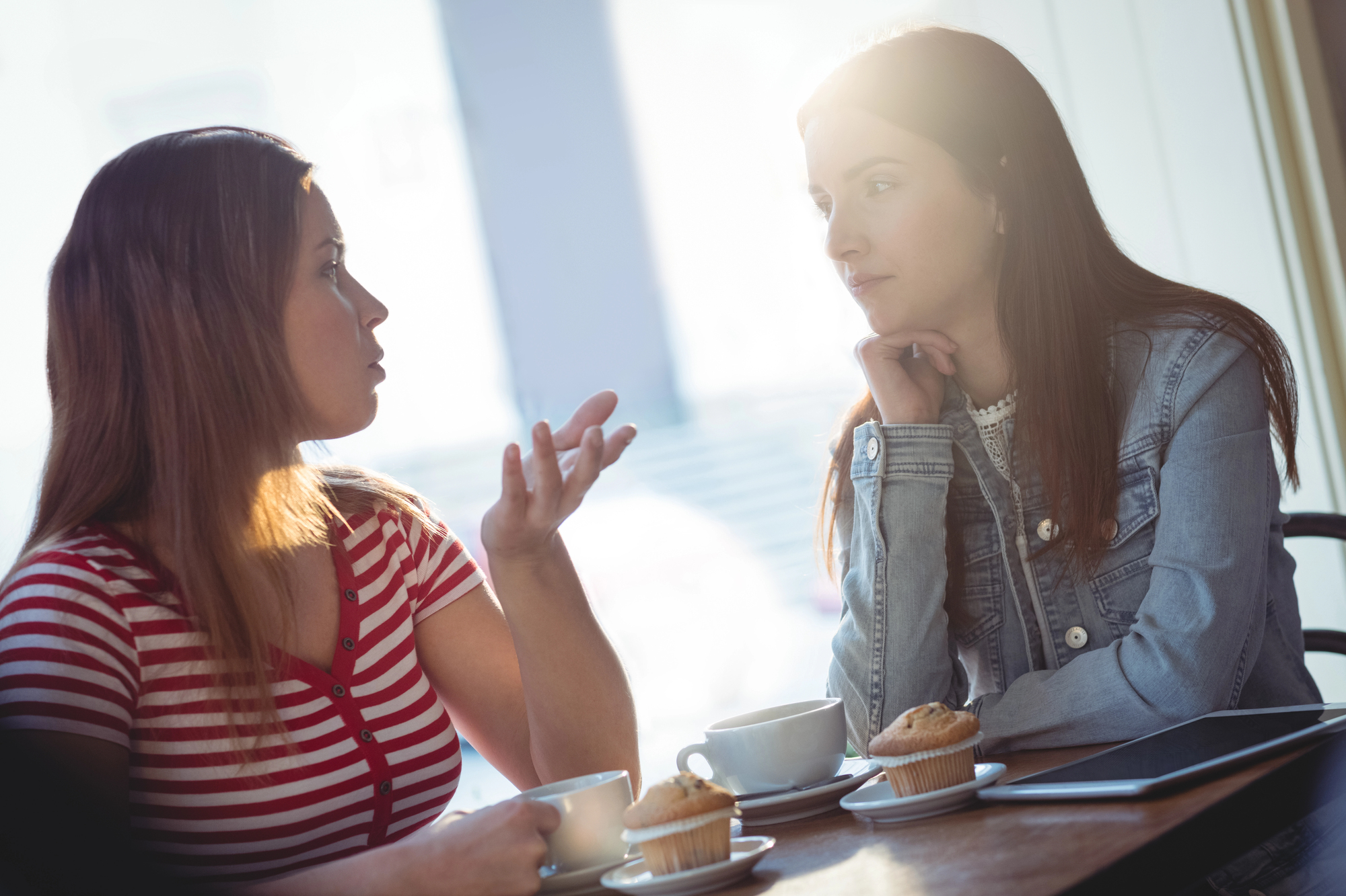 Two women are sitting at a table in a café, engaged in a conversation. One has her hand raised in a gesturing motion, while the other rests her chin on her hand, listening intently. On the table are two coffee cups, saucers, muffins, and a tablet.