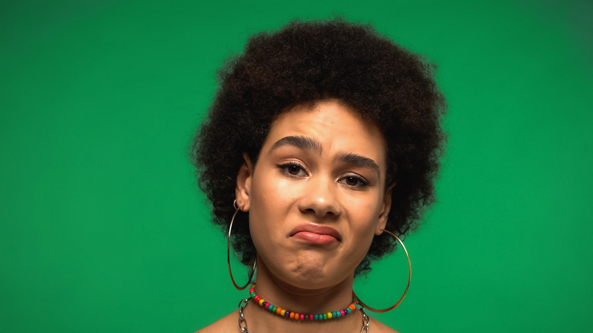 A person with an afro hairstyle is standing against a green background, wearing large hoop earrings and a colorful beaded necklace. They have a neutral expression with a slight frown.