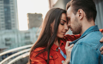 A couple stands closely on an outdoor balcony, their foreheads touching. The woman, wearing a red jacket, gently holds the collar of the man's denim jacket. Both have their eyes closed in a tender moment. City buildings and a handrail are visible in the background.