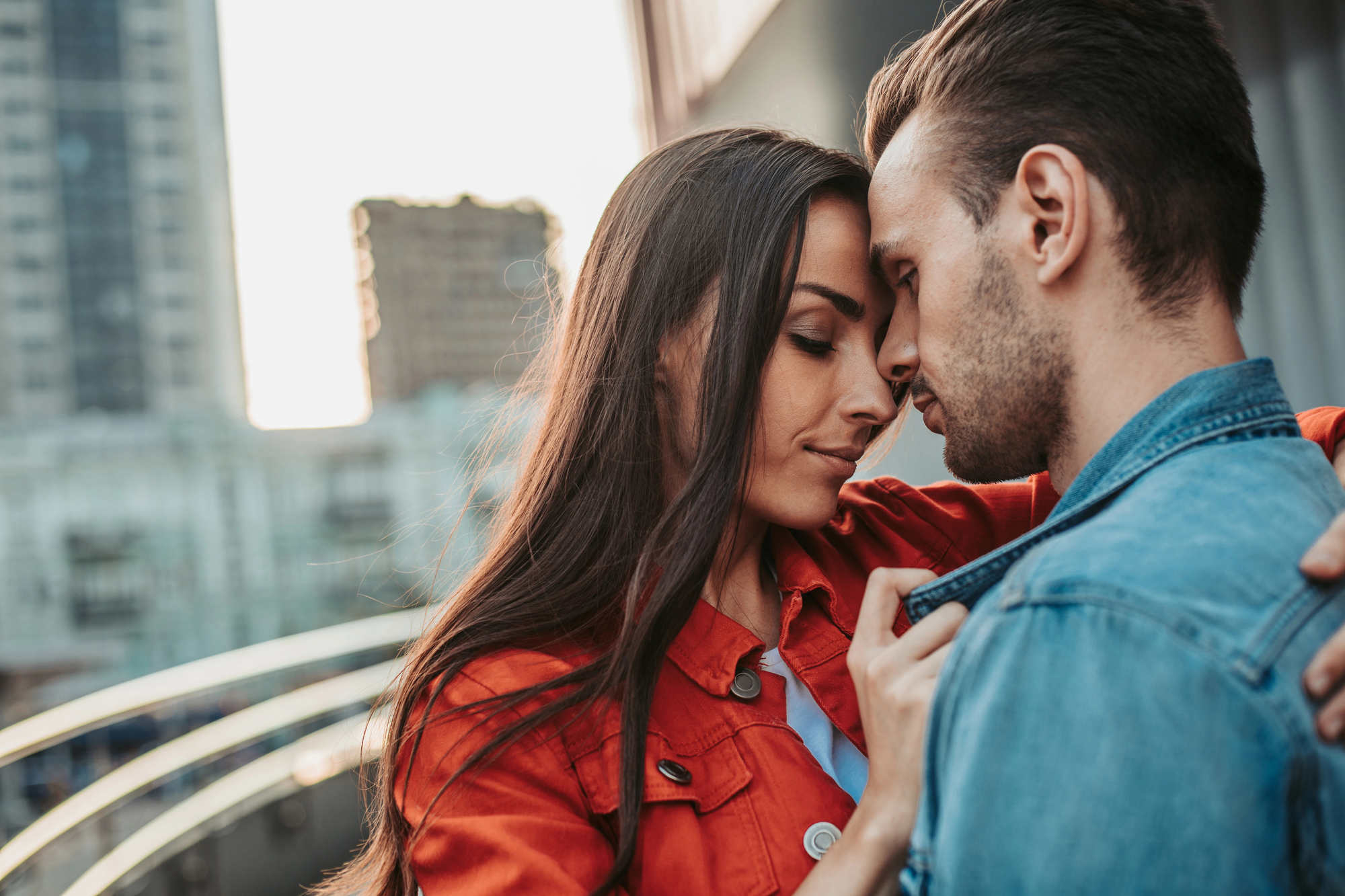 A couple stands closely on an outdoor balcony, their foreheads touching. The woman, wearing a red jacket, gently holds the collar of the man's denim jacket. Both have their eyes closed in a tender moment. City buildings and a handrail are visible in the background.