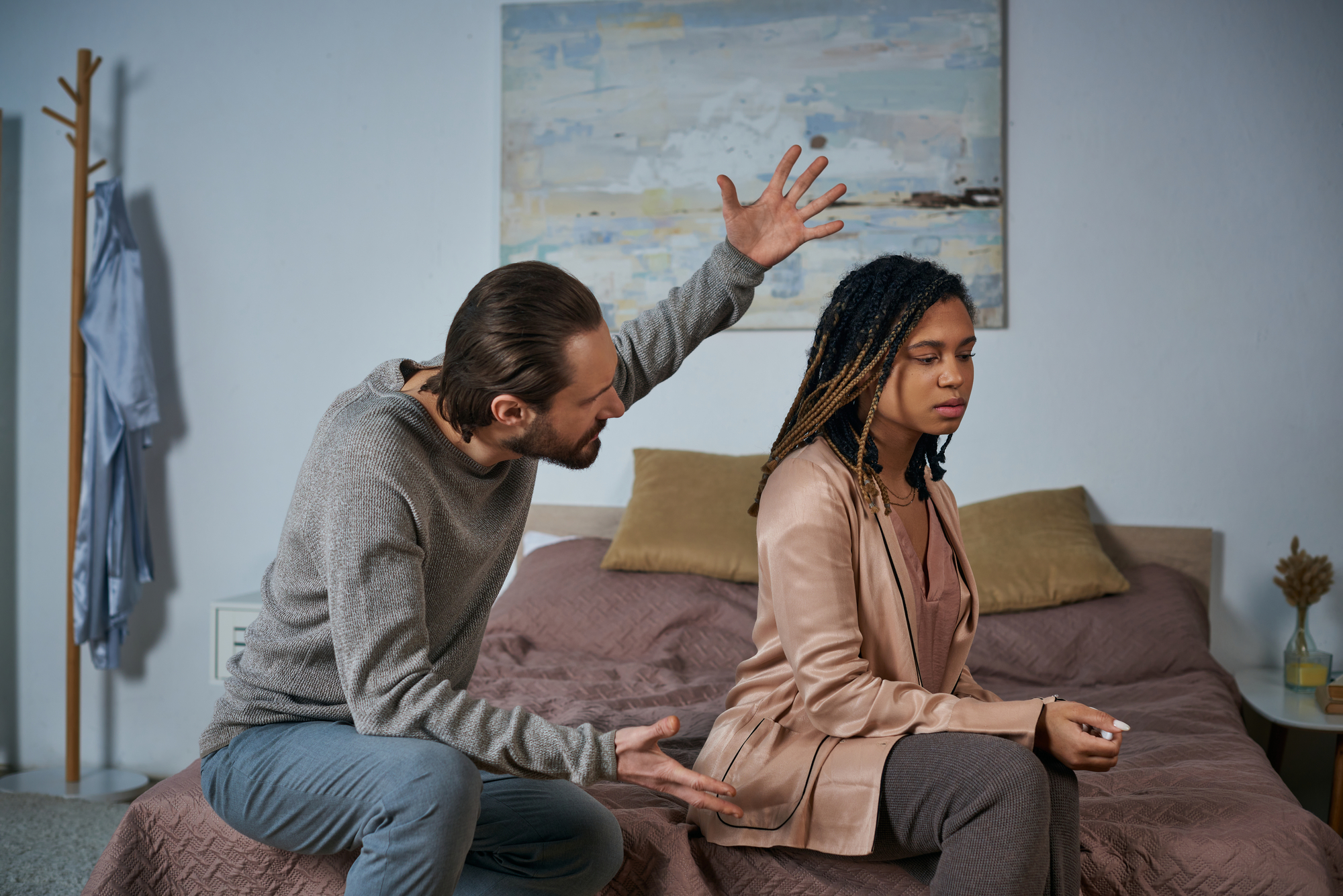 A man with a serious expression is sitting on a bed, gesturing with open hands towards a woman sitting next to him. The woman, looking away with a stoic expression, is wearing a beige jacket and has braided hair. A painting hangs on the wall behind them.