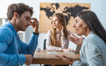 Three young professionals are having a discussion in an office. A man, looking distressed, holds his head with his hand, while a woman in front of him is speaking passionately. Another woman sits in the background looking contemplative. A world map is on the wall.