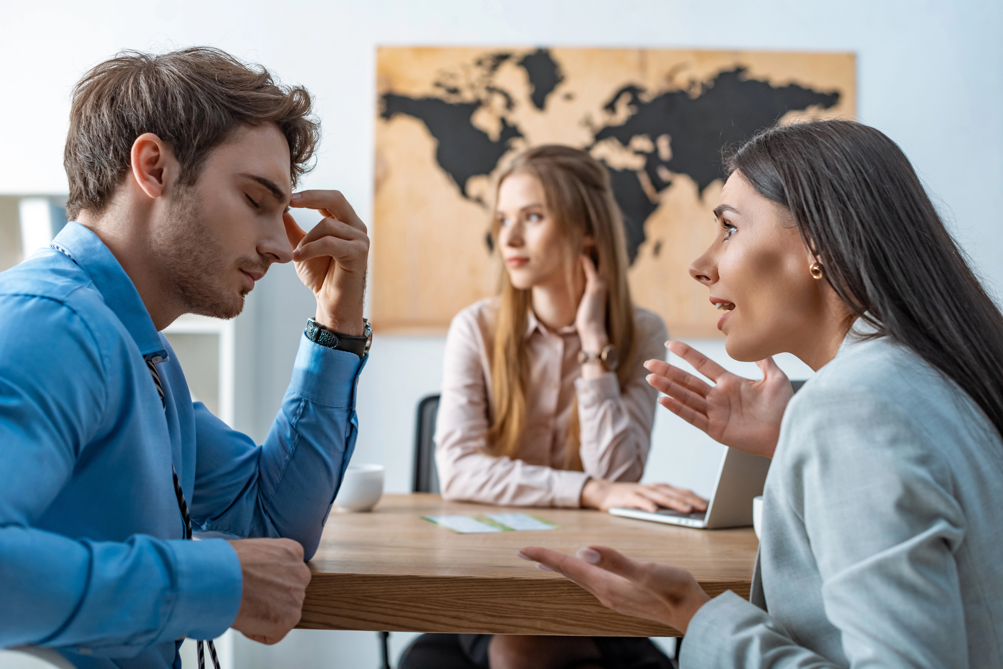 Three young professionals are having a discussion in an office. A man, looking distressed, holds his head with his hand, while a woman in front of him is speaking passionately. Another woman sits in the background looking contemplative. A world map is on the wall.