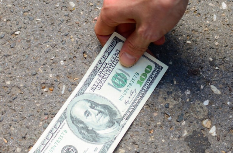 A hand is picking up a one hundred dollar bill from the ground. The bill features the portrait of Benjamin Franklin. The ground is composed of rough, textured concrete with small pebbles embedded in it.