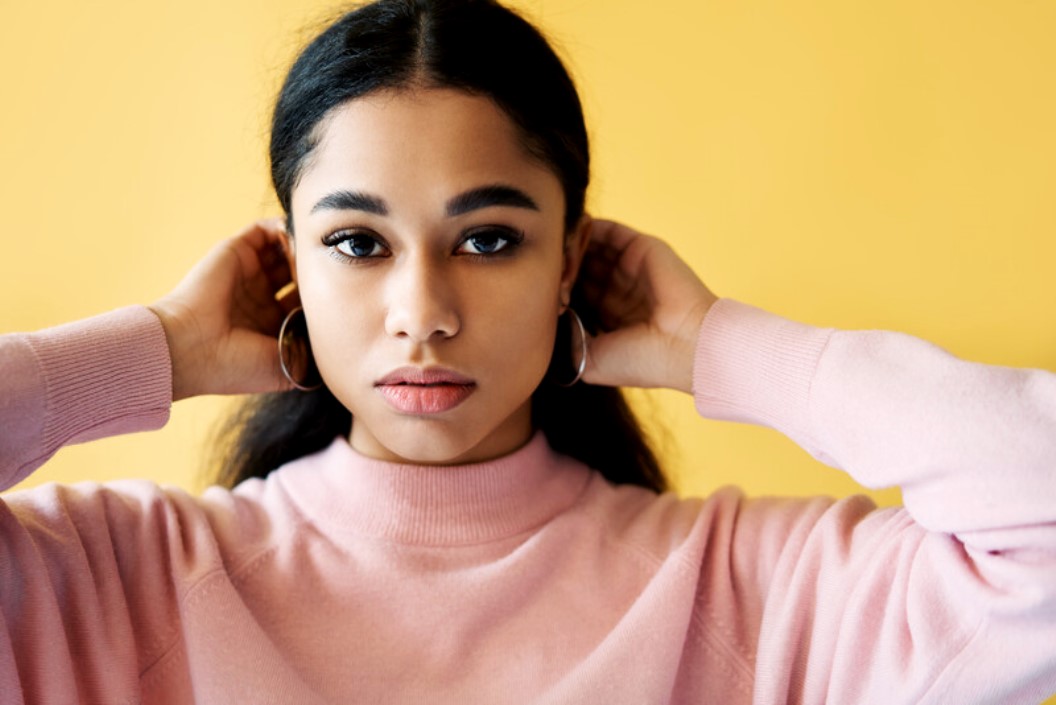 A young woman with long dark hair and hoop earrings adjusts her hair with both hands. She is wearing a pink sweater and stands against a yellow background. She looks directly at the camera with a neutral expression.