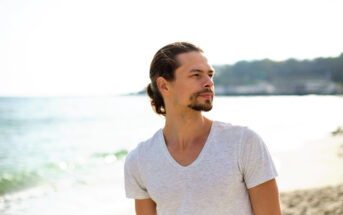 A man with long hair tied back is standing on a beach, looking off into the distance. He is wearing a light gray V-neck T-shirt. The background features a calm sea and a hazy, out-of-focus shoreline. The atmosphere is bright and sunny.