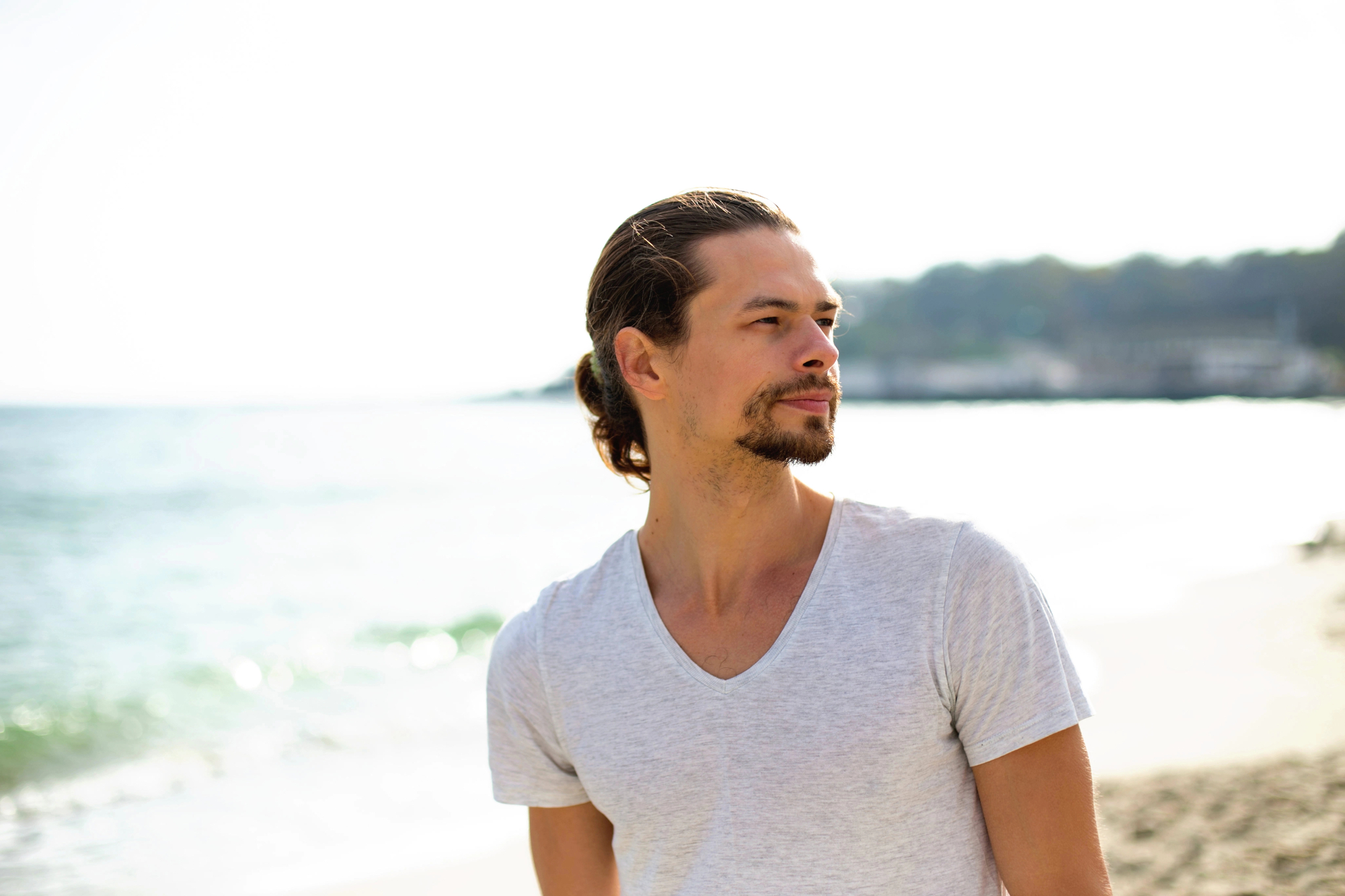 A man with long hair tied back is standing on a beach, looking off into the distance. He is wearing a light gray V-neck T-shirt. The background features a calm sea and a hazy, out-of-focus shoreline. The atmosphere is bright and sunny.