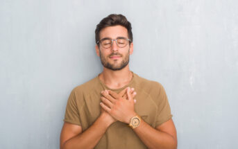 A man with short dark hair and a beard, wearing glasses and a brown t-shirt, stands against a light gray wall. He has his eyes closed and his hands crossed over his chest in a calm, meditative pose.