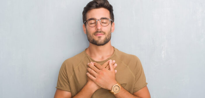 A man with short dark hair and a beard, wearing glasses and a brown t-shirt, stands against a light gray wall. He has his eyes closed and his hands crossed over his chest in a calm, meditative pose.