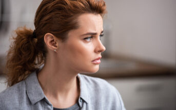 A woman with red hair tied in a ponytail is looking to the right with a serious expression. She is wearing a grey blouse and small stud earrings. The background is blurred, focusing attention on her face.