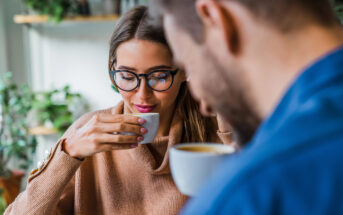 A woman with glasses, wearing a light brown sweater, sips from a white cup. A bearded man, partially visible in the foreground, also holds a white cup. The background is blurred with green plants and shelves. They appear to be in a cozy, casual setting.