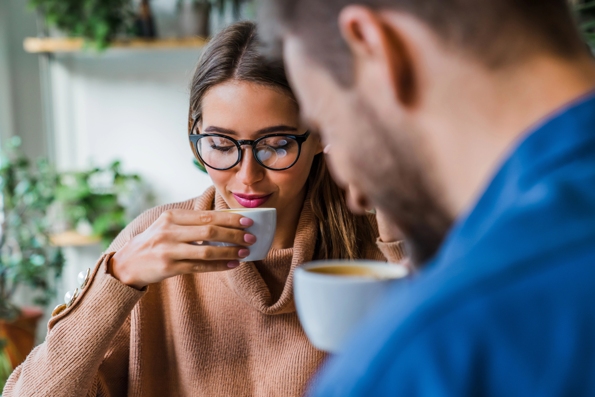 A woman with glasses, wearing a light brown sweater, sips from a white cup. A bearded man, partially visible in the foreground, also holds a white cup. The background is blurred with green plants and shelves. They appear to be in a cozy, casual setting.