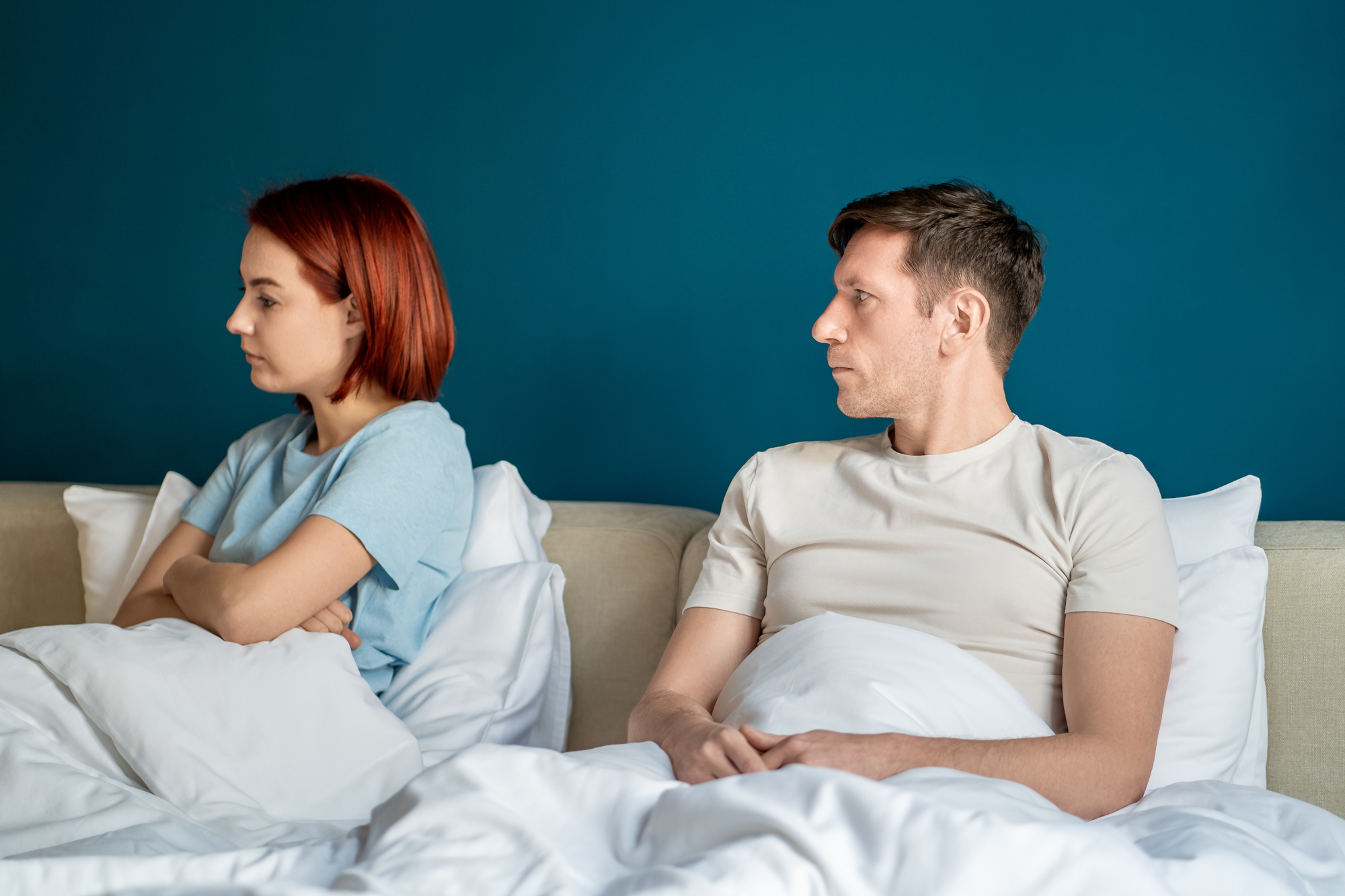 A woman with short red hair and a man with short brown hair sit in bed under white blankets, facing away from each other and looking upset. The woman sits with her arms crossed, while the man rests his arms on the bed. Both have neutral blue and white attire.