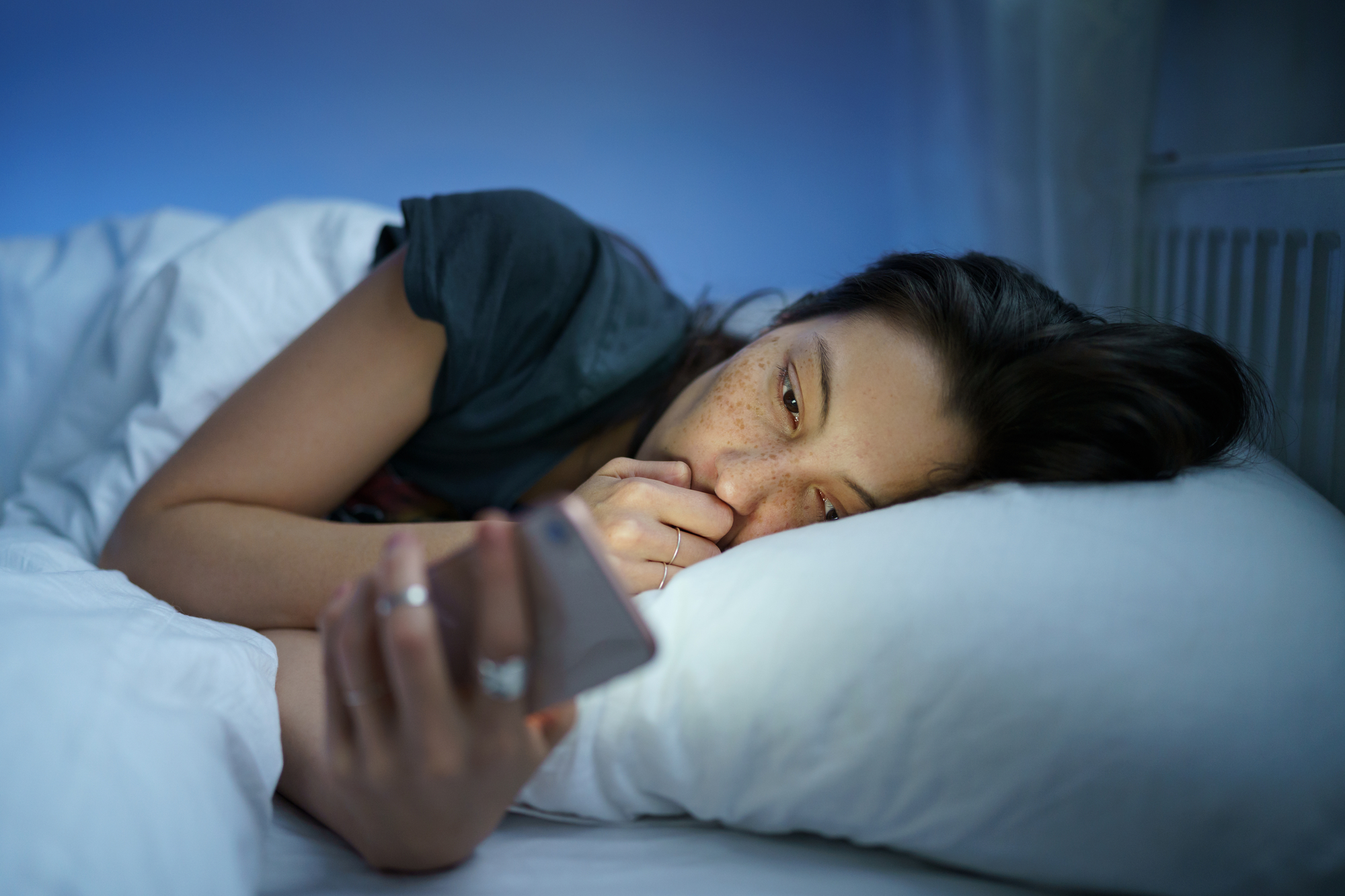 A person with long dark hair lies in bed, resting their head on a white pillow while looking at a smartphone held in their right hand. The person is under a white blanket, and the room has a soft blue light. The person has a pensive expression.