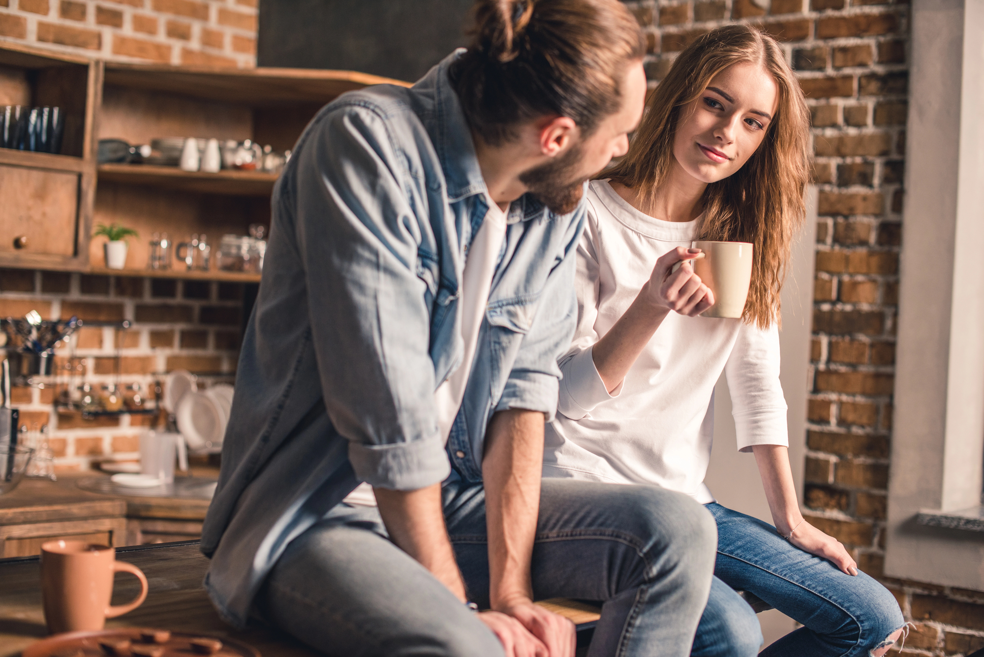 A man and a woman sit on a kitchen counter, both holding coffee mugs. The man has a beard and a bun, wearing a denim shirt, and the woman has long hair, wearing a white sweater. They are engaged in conversation with a warmly lit, rustic kitchen in the background.