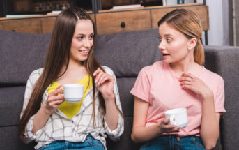 Two women are sitting on a sofa, holding white mugs, and engaged in conversation. The woman on the left has long brown hair, wears a plaid shirt and yellow top. The woman on the right has blonde hair, wears a pink shirt, and has an expression of surprise.