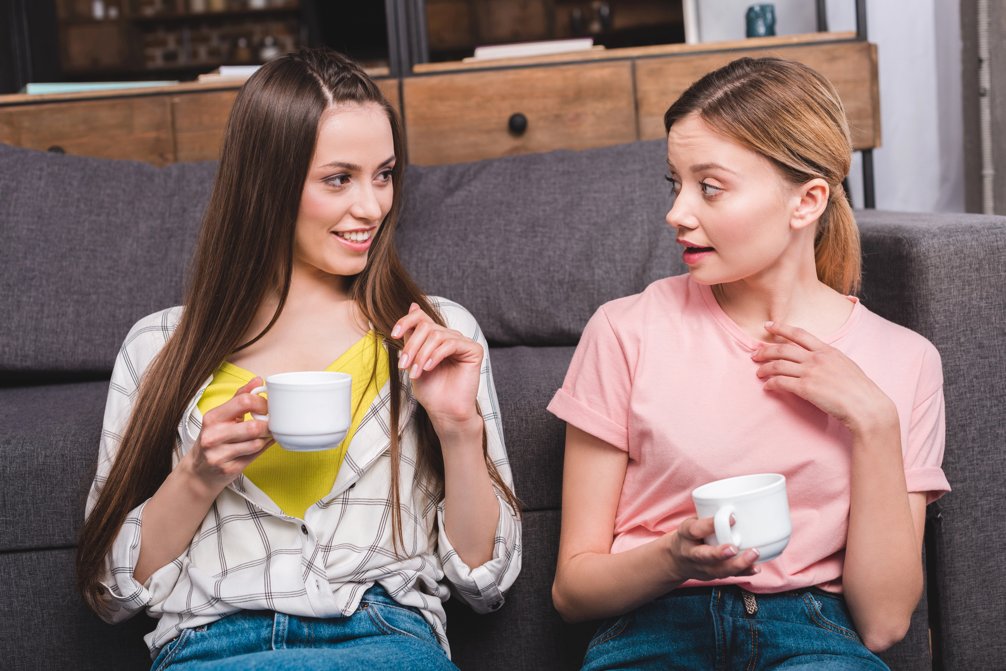 Two women are sitting on a sofa, holding white mugs, and engaged in conversation. The woman on the left has long brown hair, wears a plaid shirt and yellow top. The woman on the right has blonde hair, wears a pink shirt, and has an expression of surprise.