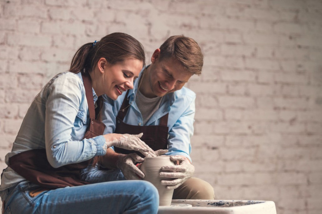 A woman and a man, both wearing aprons, are happily shaping a clay pot on a pottery wheel together. They are indoors, against a brick wall background, focused on creating the pottery piece. Their hands are covered in clay as they work collaboratively.