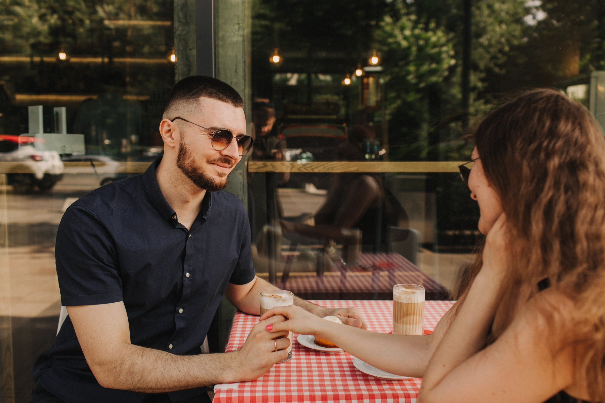 Two people sit at a table with a red checkered tablecloth outside a cafe. They are holding hands, smiling, and wearing sunglasses. Drinks, including iced coffees, are on the table. The background shows a window reflecting greenery and outdoor furniture.