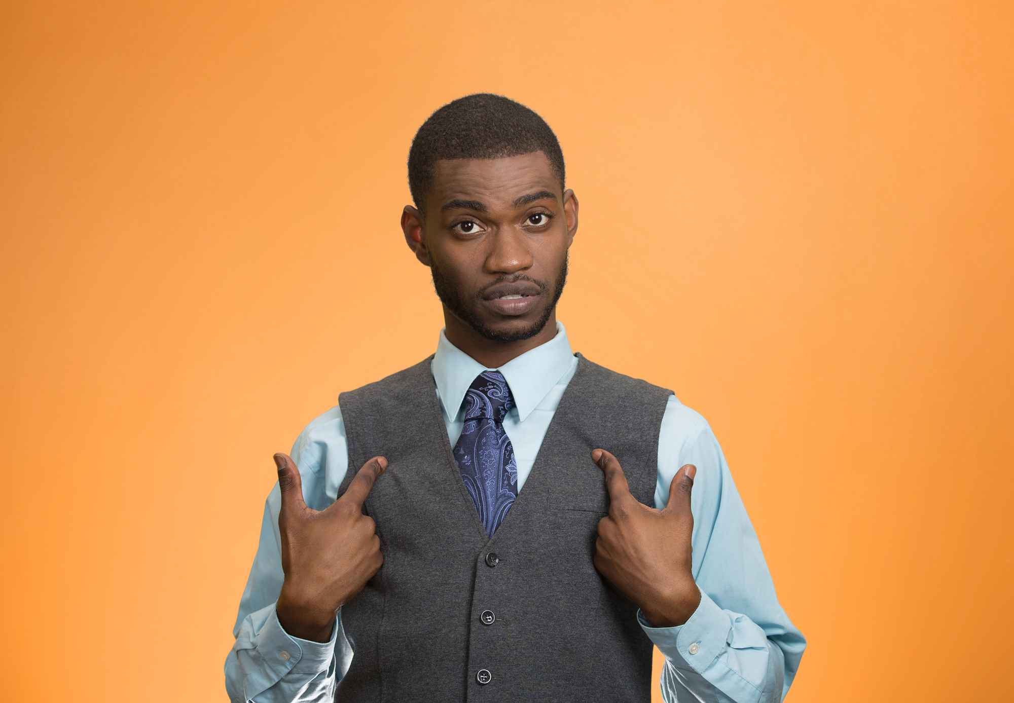 A man with a serious expression points to himself with both thumbs. He is wearing a light blue shirt, a dark gray vest, and a blue patterned tie, standing in front of an orange background.