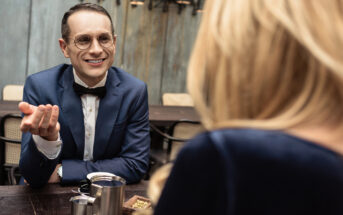 A man in a blue suit, bow tie, and glasses is sitting at a table in a restaurant, engaging in conversation with a woman with blonde hair, who is turned away from the camera. The table has a metal cup and some food items on it.