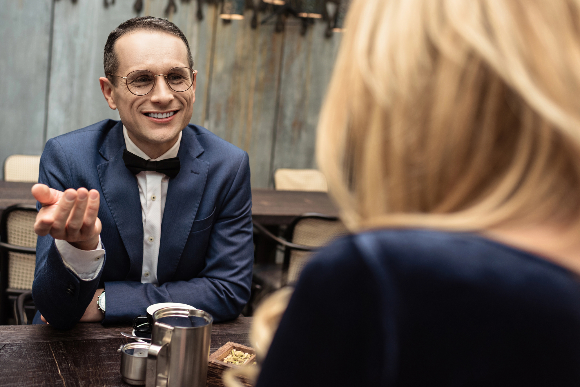 A man in a blue suit, bow tie, and glasses is sitting at a table in a restaurant, engaging in conversation with a woman with blonde hair, who is turned away from the camera. The table has a metal cup and some food items on it.