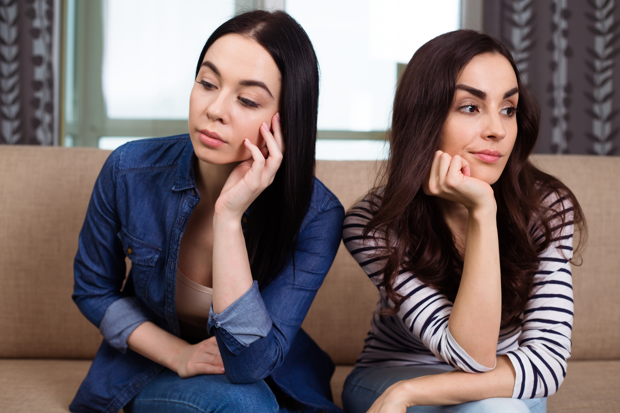 Two women with long dark hair sit on a beige couch, looking away from each other thoughtfully. One wears a denim jacket and rests her head on her hand, while the other wears a striped shirt and rests her chin on her hand with a slight smile.