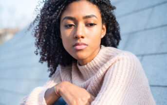 A woman with curly hair is gazing into the distance with a thoughtful expression. She is wearing a light pink sweater and appears to be seated outdoors against a background of gray stone steps.