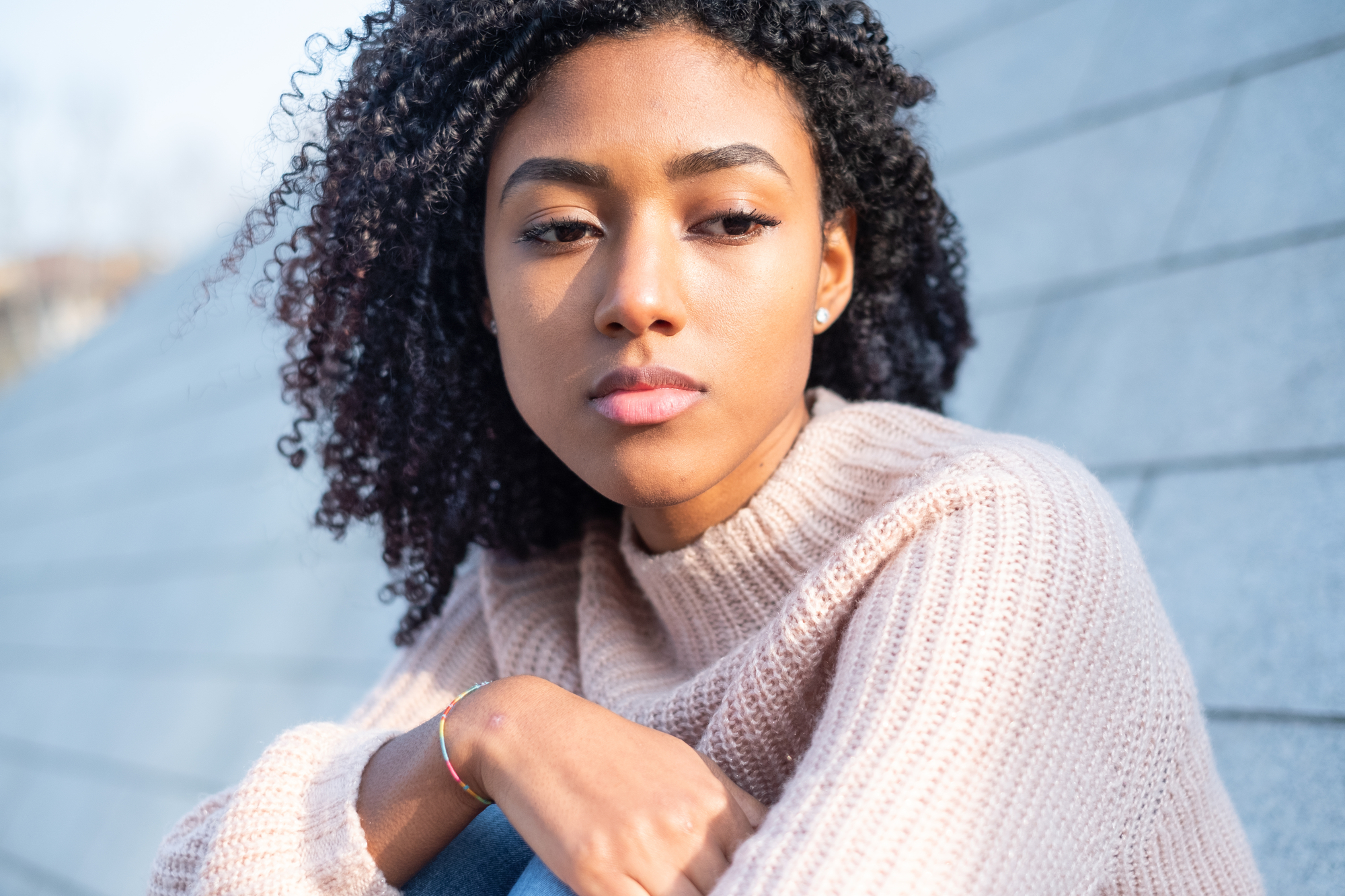 A woman with curly hair is gazing into the distance with a thoughtful expression. She is wearing a light pink sweater and appears to be seated outdoors against a background of gray stone steps.