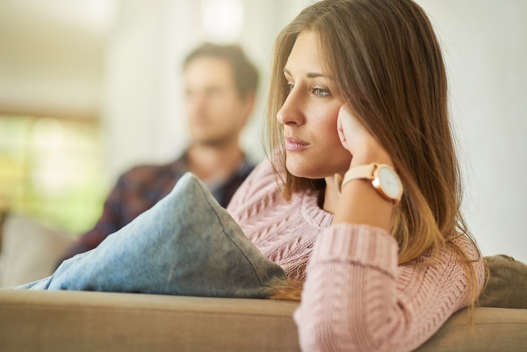 A woman with long hair, wearing a pink sweater and a watch, rests her head on her hand while sitting on a couch, looking thoughtful. A blurred person in a checkered shirt sits in the background. The scene appears to be indoors with soft lighting.