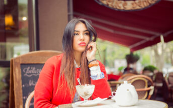 A woman with long, dark hair sits at an outdoor café table, resting her chin on her hand. She wears a red coat and looks thoughtful. In front of her is a dessert glass with whipped cream and a teapot. Her phone is on the table next to her.