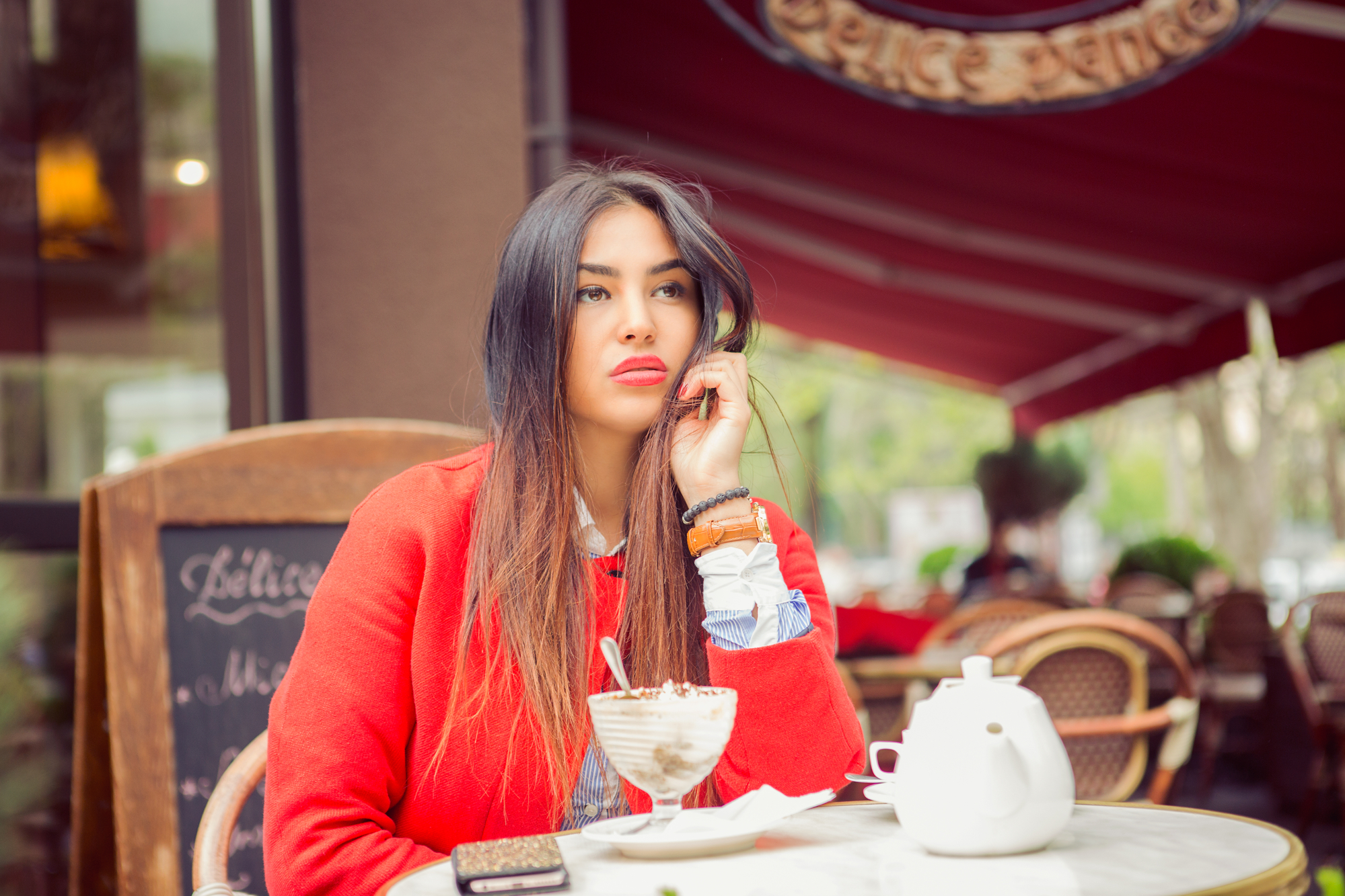 A woman with long, dark hair sits at an outdoor café table, resting her chin on her hand. She wears a red coat and looks thoughtful. In front of her is a dessert glass with whipped cream and a teapot. Her phone is on the table next to her.