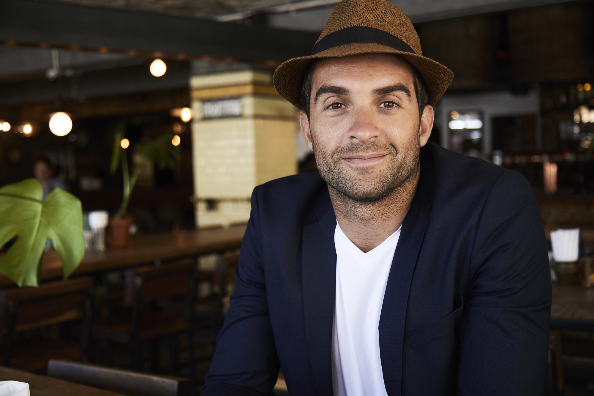 A man wearing a brown fedora and navy blazer over a white T-shirt is smiling while sitting indoors at what appears to be a rustic café or restaurant. The background shows wooden tables, chairs, and warm ambient lighting.